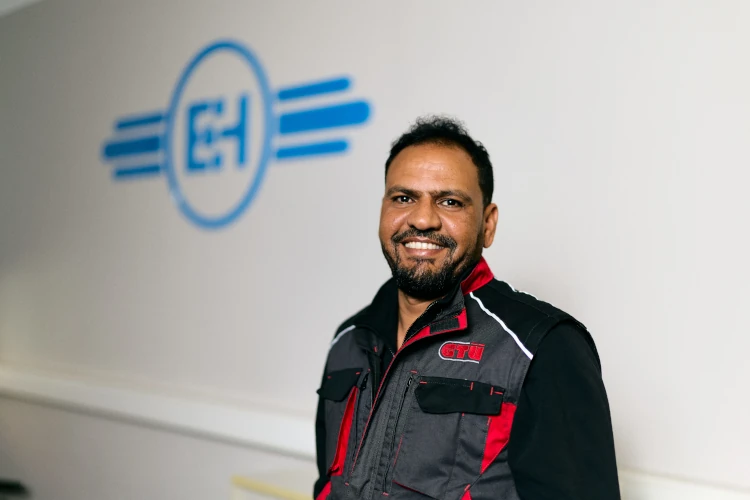 Ein lächelnder Mann in Arbeitskleidung steht vor einem Kfz-Prüfdienst-Logo an der Wand.